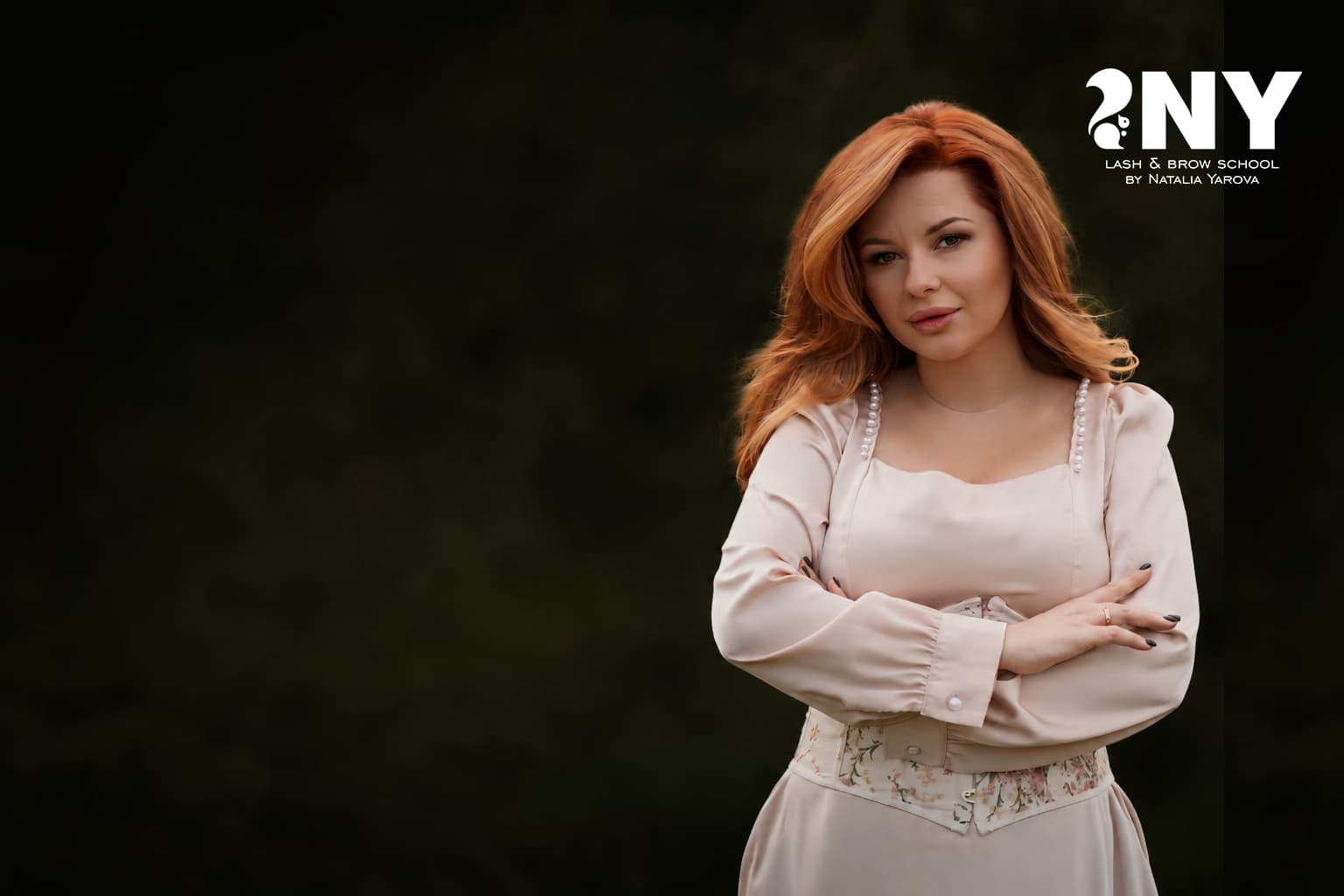 Red-haired woman in a pearl-trimmed dress posing with arms crossed against a dark background.